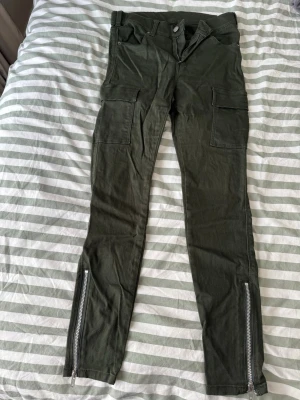 Mörkgröna cargobyxor med dragkedjor - Säljer ett par mörkgröna cargobyxor med raka ben, stora fickor på sidorna och dragkedjor vid bensluten. Byxorna har knapp och dragkedja i midjan samt bälteshällor. Perfekta för en cool och avslappnad stil.