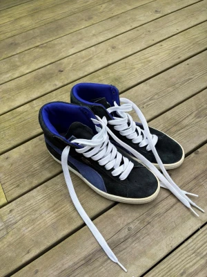 Svarta Puma Suede High Sneakers - Snygga svarta high top sneakers från Puma med blå detaljer och vit sula. Skorna är gjorda i mocka och har vita skosnören samt blått foder på insidan. Klassisk Puma-logga i guld på sidan och ikonisk formstripe i blå mocka.