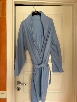 Ljusblå morgonrock med skärp - Ljusblå morgonrock med långa ärmar och tillhörande skärp i midjan. Perfekt för att chilla hemma eller efter duschen. Saknar storlek, uppskattad till S.