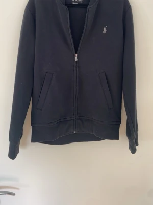 Svart zip-hoodie från Ralph Lauren - Svart hoodie med dragkedja från Ralph Lauren. Klassisk design med två fickor framtill och broderad logga på bröstet. Tillverkad i mjukt bomullsmaterial och har ribbade muddar vid ärmslut och nederkant. Perfekt för en avslappnad stil. Storlek s