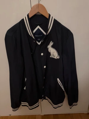 Svart bomberjacka från Gant med kanin - Svart bomberjacka från Gant med vita ränder på krage, ärmslut och nederkant. Stor kaninpatch på bröstet och GANT-tryck i vitt på ryggen. Jackan har klassisk bomberstil med tryckknappar och fickor. Perfekt för en sportig och trendig look.