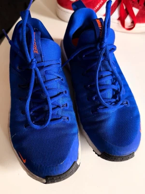 Blå Nike sneakers med vit sula - Snygga blå sneakers från Nike med vit sula och svarta detaljer. Skorna har klassisk låg modell, blåa skosnören och orange Nike-logga på tungan och sidan. Ovandelen är i textil för en sportig och luftig känsla. Perfekta för en aktiv vardag. Nyskick då dessa vart bara provade.
