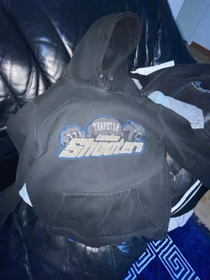 Trapstar London Shooters hoodie - Svart hoodie från Trapstar med stor broderad logga och texten 'London Shooters' på bröstet. Hoodien har huva och magficka, samt coolt tryck i blått, grått och vitt. Perfekt för dig som gillar streetwear och vill sticka ut.