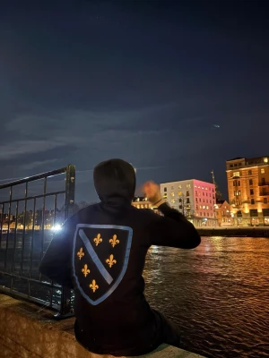 Bosnia tröja  - Säljer en svart hoodie med stor blå vapensköld på ryggen, dekorerad med gula liljor och ett diagonalt blått band. Hoodien har huva och en avslappnad passform, perfekt för dig som vill sticka ut med en unik design.