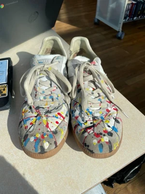 Maison Margiela sneakers med färgstänk - Unika sneakers från Maison Margiela i vitt och beige läder och mocka, dekorerade med färgstänk i rött, blått, gult och svart. Klassisk låg modell med gummisula och beige snörning. Perfekta för dig som vill sticka ut med din stil. Defekt på sista bild!
