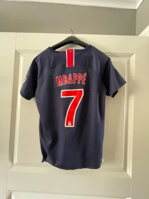 PSG Mbappé matchtröja Nike - Mörkblå PSG matchtröja med Mbappé och nummer 7 på ryggen. Framsidan har Fly Emirates-logga, Nike-märke och PSG-emblem. Röd och vit detalj i mitten. Perfekt för fotbollsfans som vill visa sitt stöd för Paris Saint-Germain.