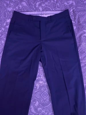 Mörkblå kostymbyxor - Snygga mörkblå kostymbyxor med klassiskt snitt och pressveck. Byxorna har hällor för bälte, knapp och dragkedja fram samt en bakficka med knapp. Perfekta för en stilren look.