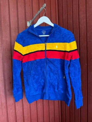 Färgglad ziptröja från Polo Ralph Lauren - Säljer en blå ziptröja från Polo Ralph Lauren med färgglada ränder i gult, rött och svart över bröstet. Tröjan har ståkrage, dragkedja framtill och ribbade muddar. Perfekt för dig som gillar sportig och retro stil. Barn storlek L! Passar dam xs/s 