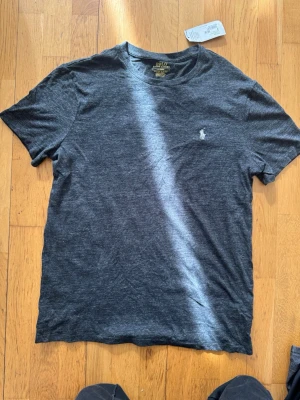 Mörkgrå t-shirt från Polo Ralph Lauren - Helt ny oanvänd snygg  mörkgrå t-shirt från Polo Ralph Lauren med klassisk rund hals och broderad vit logga på bröstet. Modellen är custom slim fit och materialet är mjuk bomullsmix som känns skön mot huden. Perfekt för en stilren och avslappnad look.