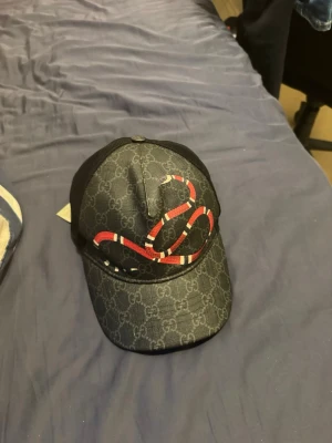 Gucci keps med orm-motiv - Snygg svart keps från Gucci med klassiskt GG-mönster och en röd-vit orm broderad framtill. Baksidan är i mesh-material med justerbart spänne och en röd detalj. Perfekt för dig som vill sticka ut med en lyxig accessoar. Skick 6.5/10 köpt i london år 2020 säljer eftersom den passar ej min stil!