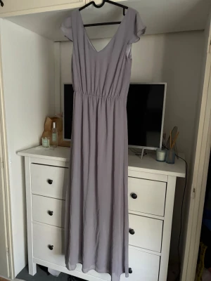 Ljuslila långklänning med v-ringning - Långklänning i ljuslila med v-ringad hals och korta ärmar. Klänningen har ett elastiskt band i midjan som ger en snygg passform och är tillverkad i ett lätt och flödande material. Perfekt för dig som gillar en enkel och stilren look. Där fram har den en skada men inget som är jätte synligt och går nog enkelt att fixa om man vill