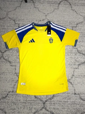 Sverige vm matchtröja Adidas gul/blå - Säljer en  Sverige vm matchtröja från Adidas i gult med blå detaljer och vita ränder på axlarna. Tröjan har broderat landslagsemblem på bröstet och är tillverkad i ett lätt, ventilerande funktionsmaterial. Perfekt för fotboll eller träning.