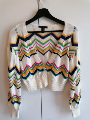 Mönstrad stickad kofta från Forever 21 - Färgglad stickad kofta i kort modell från Forever 21 med zigzag-mönster i blått, gult, grönt, rosa och vitt. Koftan har v-ringning, knappar framtill och ribbade muddar. Perfekt för dig som gillar retrovibbar och vill sticka ut lite extra.