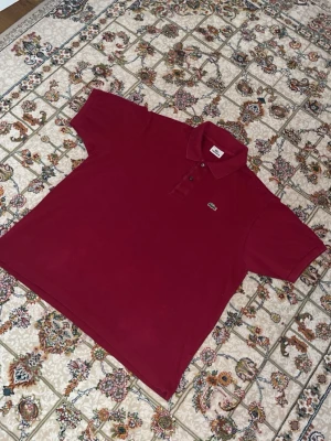 Vinröd pikétröja från Lacoste - Vinröd pikétröja från Lacoste med klassisk krage och knappslå framtill. Tröjan har korta ärmar och den ikoniska krokodilloggan broderad på bröstet. Perfekt för en stilren och avslappnad look.