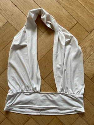 Vit halterneck linnetopp - Snygg vit halterneck linnetopp med djup urringning och bred resår nedtill. Toppen är ärmlös och tillverkad i ett mjukt, stretchigt material som känns skönt mot huden. Perfekt att styla med jeans eller kjol för en trendig look.