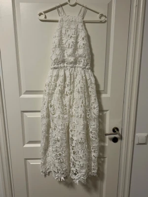 Vit långklänning med spetsdetaljer - Superfin vit långklänning med blommig spets och tunna axelband som korsas i ryggen. Klänningen har öppen rygg och ett elegant fall. Perfekt för studenten 