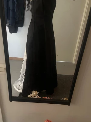 Vacker balklänning  - Elegant svart långklänning med vit panel på baksidan som har ett vackert blommigt mönster. Klänningen har korset i ryggen och är ärmlös. Jag har köpt den begagnade men den ser helt ny ut förutom att det saknas några stenar från mönstret på vissa ställen men det är inget man lägger märke till. Den är lång på mig även när jag har högklackat och jag 165 cm. Den passar väldigt bra som en balklänning. Den är storlek 11/12.