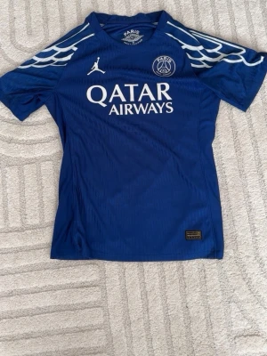 Blå PSG matchtröja Jordan x Nike - Snygg blå PSG matchtröja med vita detaljer och Qatar Airways-tryck på bröstet. Nummer 10 och O. Dembélé på ryggen samt Champions League-märke på ärmen. Tillverkad i lätt och ventilerande material, perfekt för fotboll eller träning.