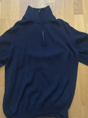 Marinblå stickad tröja Navigare - Mörkblå stickad tröja från Sail Racing med halv dragkedja och hög krage. Insidan av kragen är röd för en snygg kontrast. Tröjan har ribbstickade muddar och är perfekt för lager-på-lager. Klassisk och stilren design.