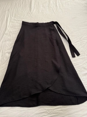 Svart omlottkjol från vero moda  - Stilren svart omlottkjol  med knytband i midjan och dragkjedia. Kjolen är lång och har en enkel, elegant design som passar perfekt till många olika looks.