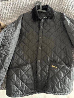 Svart quiltad jacka från Barbour - Quiltad svart jacka från Barbour med klassisk krage i manchester och tryckknappar framtill. Jackan har två stora fickor med Barbour-logga och ett rutmönster över hela ytan. Perfekt för höst och vår, med långärmad passform och snygg, tidlös design.