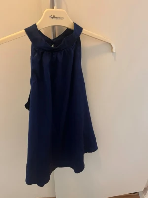 Mörkblå ärmlös halterneck-top - Snygg mörkblå halterneck-top med hög krage och knäppning i nacken. Toppen är ärmlös och har en lätt glansig finish som ger en stilren look, satin. Perfekt att matcha med jeans eller kjol för en trendig outfit. Ny och aldrig använt. 