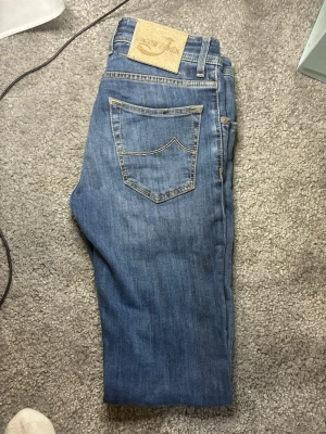 Blå jeans från Jacob Cohen - Säljer ett par klassiska blå jeans från Jacob Cohen med snygga kontrastsömmar och en patch i mockaimitation bak. Jeansen har rak passform och fem fickor. Perfekta för dig som gillar stilrena och tidlösa plagg.