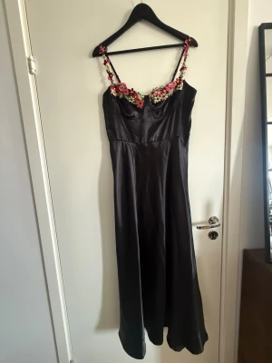 Svart långklänning med blommiga detaljer - Elegant svart långklänning i satinliknande material med tunna axelband dekorerade med broderade blommor i rosa, rött och grönt. Klänningen har en figurnära överdel och faller snyggt längs kroppen. Perfekt för dig som vill sticka ut med unika detaljer.