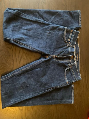 Mörkblå jeans från Nudie Jeans - Snygga mörkblå jeans från Nudie Jeans med klassisk femficksdesign och kontrastsömmar. Jeansen har raka ben och en läderpatch med logga bak i midjan. Perfekta för en stilren och avslappnad look.