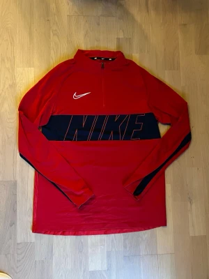 Röd långärmad Nike half zip topp - Snygg röd långärmad topp från Nike med svart panel och stor logga över bröstet. Tröjan har en half zip-dragkedja framtill och är tillverkad i ett mjukt syntetmaterial som passar perfekt till träning eller chill. Ikonisk Swoosh-detalj på bröstet och sportig look.