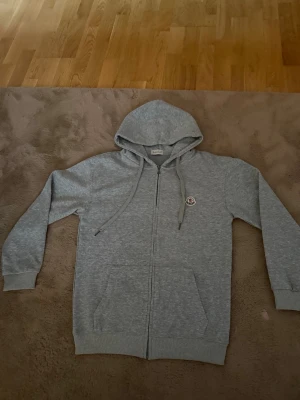 Grå Moncler kofta (size S) - Snygg grå hoodie med dragkedja från Moncler. Klassisk modell med huva och dragsnören, två fickor framtill och Moncler-logga på bröstet. Perfekt för en avslappnad och stilren look. Mjuk bomullsblandning för extra komfort. Sälja på grund av fel storlek.