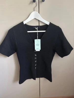Svart v-ringad topp med knappar - Stilren svart topp från Gina Tricot med v-ringning och korta ärmar. Toppen har en figurnära passform och dekorativa knappar framtill. Perfekt att styla med jeans eller kjol för en trendig look.