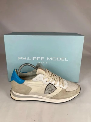 Philippe Model sneakers  - Snygga sneakers från Philippe Model i beige och vitt med detaljer i blått och silver. Skorna har mesh och mocka på ovandelen, samt en ikonisk sköldlogga på sidan. Perfekta för dig som gillar stilrena och trendiga sneakers med unika detaljer. Storlek 41 men passar 42. 