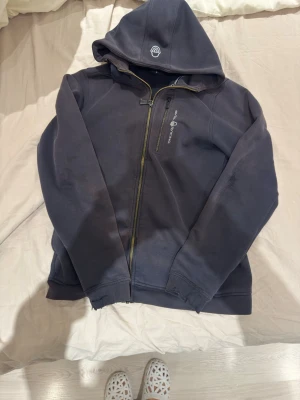 Svart hoodie från Sail Racing - Mörkblå hoodie med dragkedja från Sail Racing. Har huva med logga, bröstficka med dragkedja och broderad text på bröstet. Tillverkad i mjukt material, perfekt för en avslappnad stil. Ganska sliten med hål vid armen och blivit lite röd från solen.bilderna visar skadorna 
