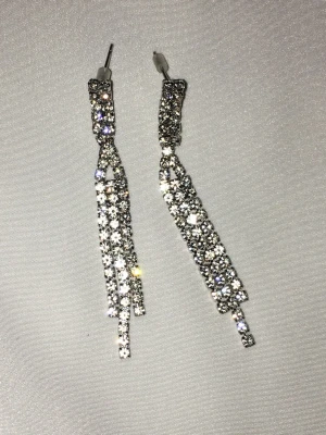 Glittriga strassörhängen - Ett par vackert gnistrande örhängen i silverfärg med flera rader av gnistrande stenar. Örhängena har en lång, hängande design som ger en lyxig och elegant känsla. 