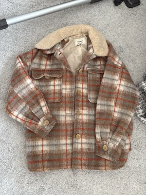 Rutig overshirt från Pull&Bear - Snygg rutig overshirt från Pull&Bear i beige, brunt och orange. Fodrad med mjuk teddykrage, två bröstfickor med lock och knappar framtill. Perfekt lager-på-lager-plagg med avslappnad passform och klassisk skjortkrage.