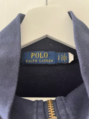 Marinblå ziptröja från Polo Ralph Lauren - Snygg marinblå ziptröja från Polo Ralph Lauren med guldfärgad dragkedja och klassisk röd logga broderad på bröstet. Tröjan har hög krage och är tillverkad i mjukt bomullsmaterial. Perfekt för en avslappnad och stilren look.
