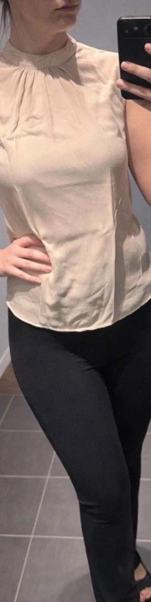 Beige ärmlös blus med hög hals - Stilren beige blus med hög hals och lätt rynk vid halsringningen. Ärmlös modell i ett tunt, mjukt material som ger en elegant känsla. Perfekt att matcha med både jeans och kjol för en clean look.