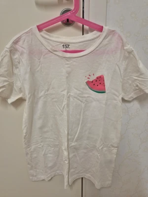 Vit t-shirt med vattenmelontryck 157 XS - Vit t-shirt från 157 i storlek XS med ett litet tryck av en vattenmelon på bröstet. Klassisk rund halsringning och korta ärmar. Perfekt för en chill och lekfull stil.