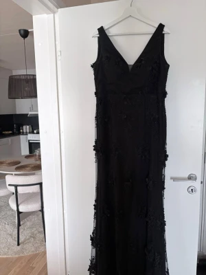 Svart lång klänning med spetsdetaljer - Elegant svart långklänning med v-ringning och vackra spetsdetaljer samt broderade blommor. Klänningen är ärmlös och har en figurnära passform, perfekt för speciella tillfällen.