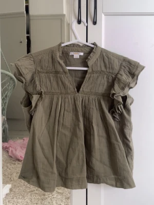 Olivgrön blus från Gina Tricot - Snygg olivgrön blus från Gina Tricot med volangdetaljer vid ärmarna och v-ringning. Blusen har en avslappnad passform och fina rynkade partier upptill. Perfekt att styla med jeans eller kjol.