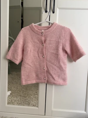 Ljusrosa kofta med pärlknappar - Söt ljusrosa kofta med korta ärmar och rund halsringning. Koftan har pärlknappar framtill och är stickad i ett mjukt material. Perfekt för lager-på-lager och en gullig touch till din outfit.