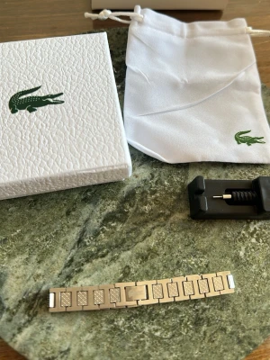 Silverfärgad klocklänk från Lacoste - Stilren silverfärgad klocklänk från Lacoste med klassisk krokodil-logga. Länken är i metall och har ett elegant rutmönster på länkarna. Levereras med original ask, tygpåse och verktyg för att justera längden.