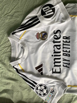 Real Madrid Mbappé matchtröja - Säljer en vit Real Madrid matchtröja från Adidas med Mbappé och nummer 10 på ryggen. Tröjan har svarta detaljer, klubbmärke, sponsorloggor och Champions League-märke på ärmen. Tillverkad i funktionsmaterial, perfekt för fotboll eller träning.
