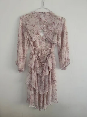 Blommig klänning med volanger - Säljer denna fina klänningen i ljusa pastellfärger med blommigt mönster (färgerna framkommer bäst på andra bilden). Klänningen har volangdetaljer, snörning och öppen rygg. Kan vara den perfekta midsommar klänningen och har länge vart slutsåld på nakds hemsida⭐️