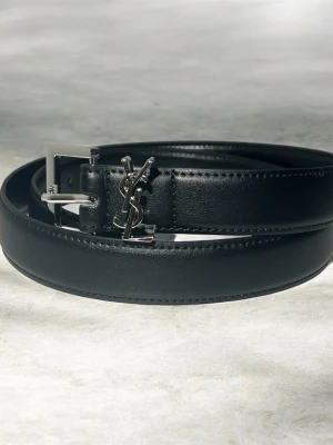 Svart skärp från Saint Laurent - Stilrent svart skärp från Saint Laurent i skinn med silverfärgat YSL-spänne. Bältet har en klassisk design med diskreta sömmar och en elegant metallspänne som ger en lyxig känsla.