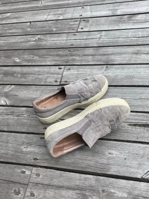 Gråa slip-on sneakers från Dasia - Gråa slip-on sneakers från Dasia med fransdetaljer på ovandelen och vit sula. Skorna är i mockaliknande material och har en låg profil som gör dem enkla att ta på. Perfekta för en avslappnad och trendig look. Stl 37 Dam. Kom med pris förslag! 