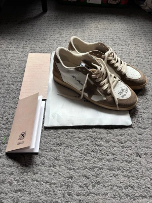 GGDB beige och vita sneakers - Snygga sneakers från Golden Goose i beige mocka och vitt läder med handskrivna detaljer på tån och sidan. Skorna har klassisk låg siluett, perforerad tå och vita snören. Märkets GGDB-logga syns på plösen. Perfekta för en trendig streetstyle-look.