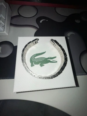 Silverfärgad klocklänk från Lacoste - Säljer en stilren silverfärgad klocklänk från Lacoste i metall. Länken har klassisk design med detaljerade länkar och den ikoniska krokodilloggan på spännet. Perfekt för dig som vill byta ut eller uppgradera din klocka med en snygg accessoar.