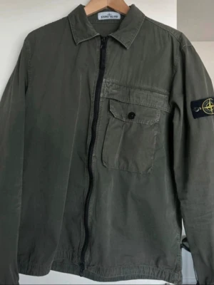 Stone Island grön overshirt - Snygg grön overshirt från Stone Island med dragkedja framtill, bröstficka med lock och knapp samt den klassiska loggan på vänster ärm. Tillverkad i bomull med avslappnad passform och krage. Perfekt lager-på-lager-plagg för en stilren look.